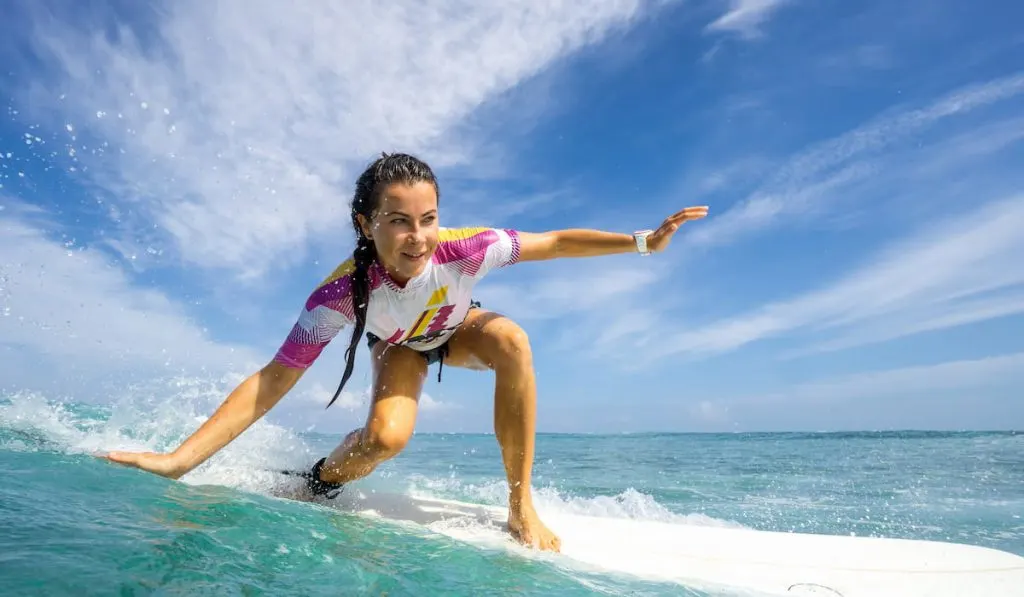 Beautiful young brunette woman in a bright bikini surfing, on the background of blue sky, clouds and transparent waves 