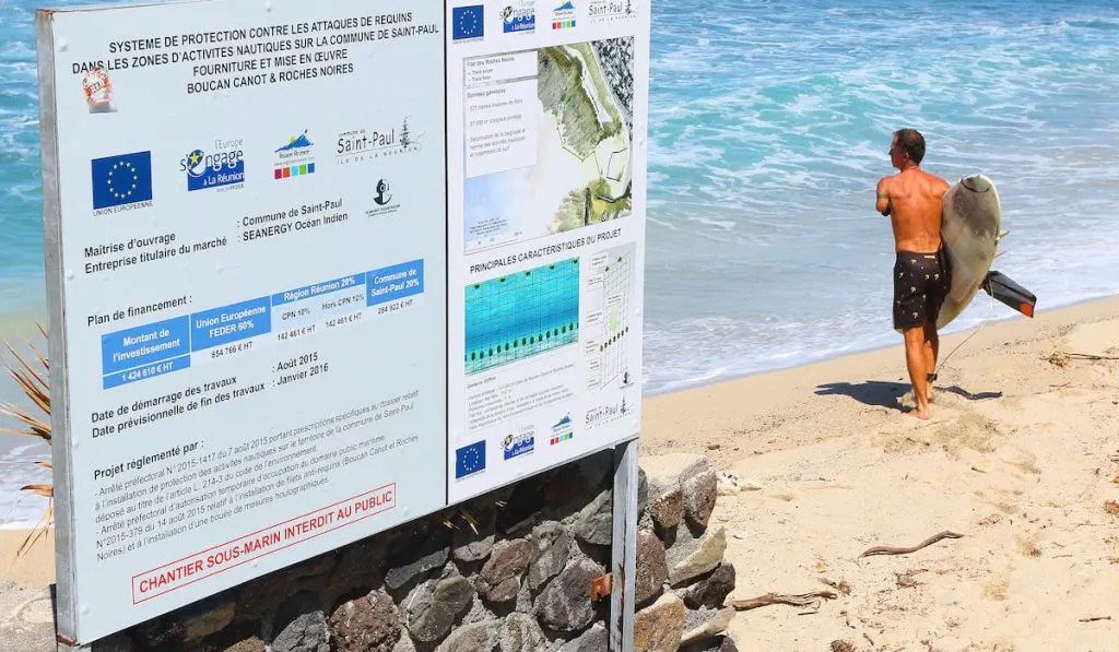 Wounded one hand surfer with damaged surfboard after shark attack standing near notice board