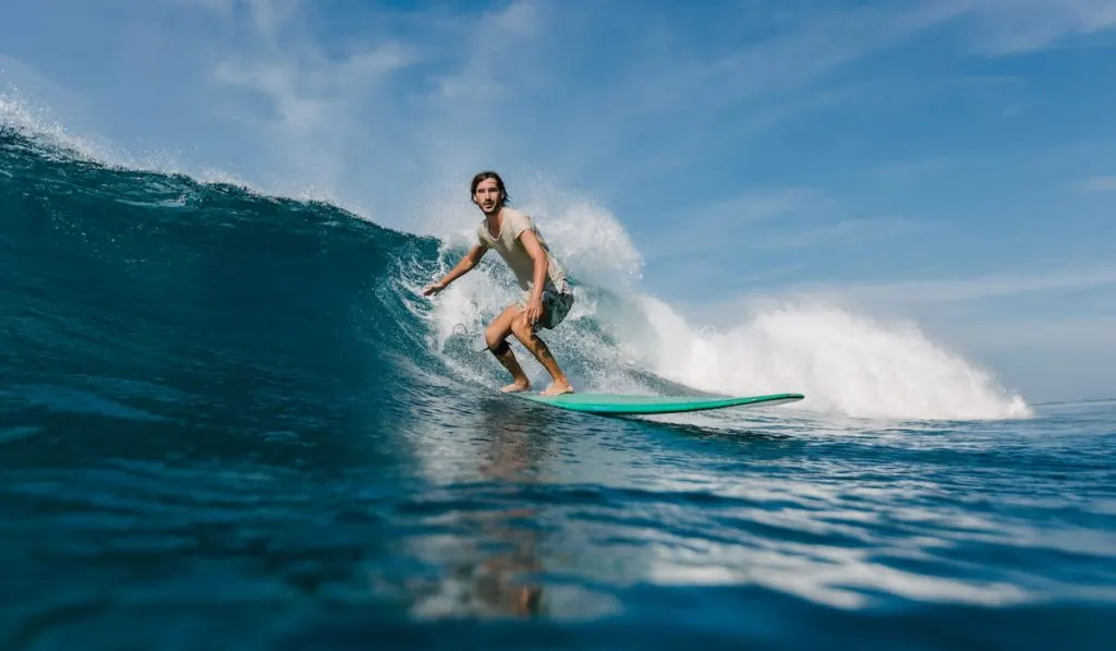 bearded young man in wet t-shirt riding waves on surfboard on sunny day