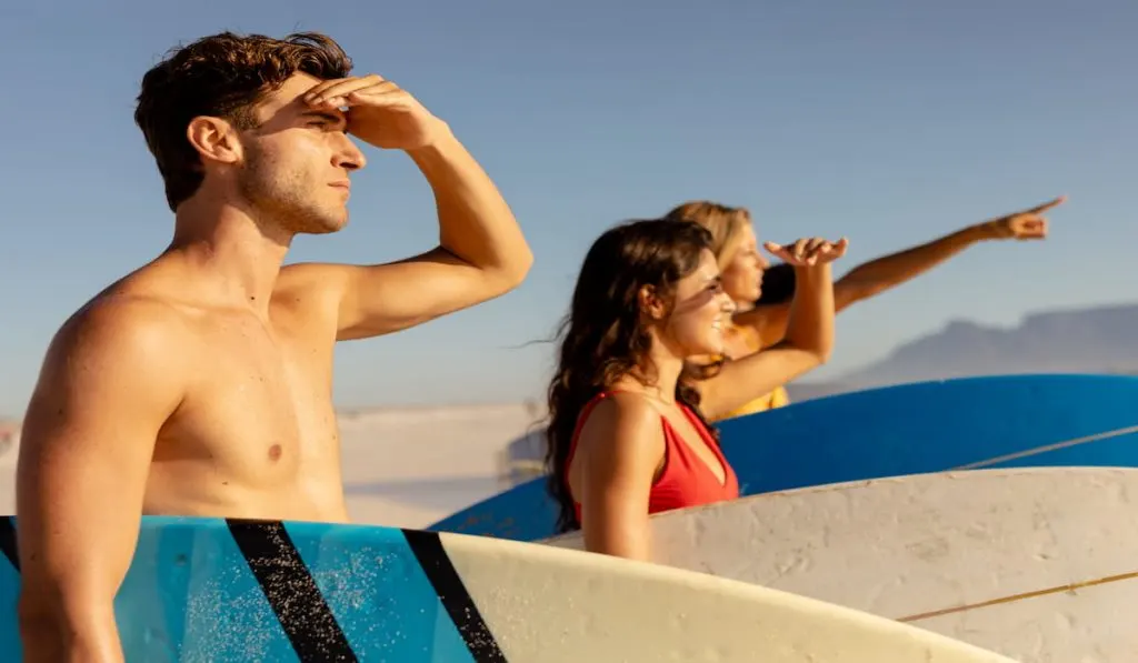 friends on a beach in the sun holding surfboards looking out to sea waiting for some waves to surf
