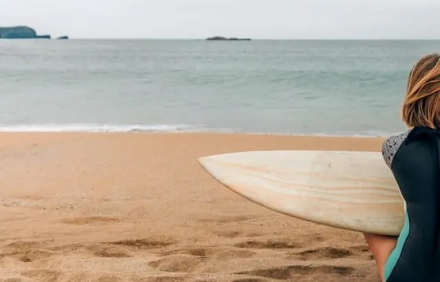 Surfer woman with wetsuit and surfboard sitting on the sand looking aside on the beach - ee220916