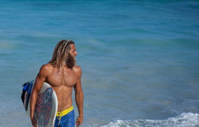 Horizontal portrait of attractive bearded curly wet man in blue shorts with surfboard - ss221208