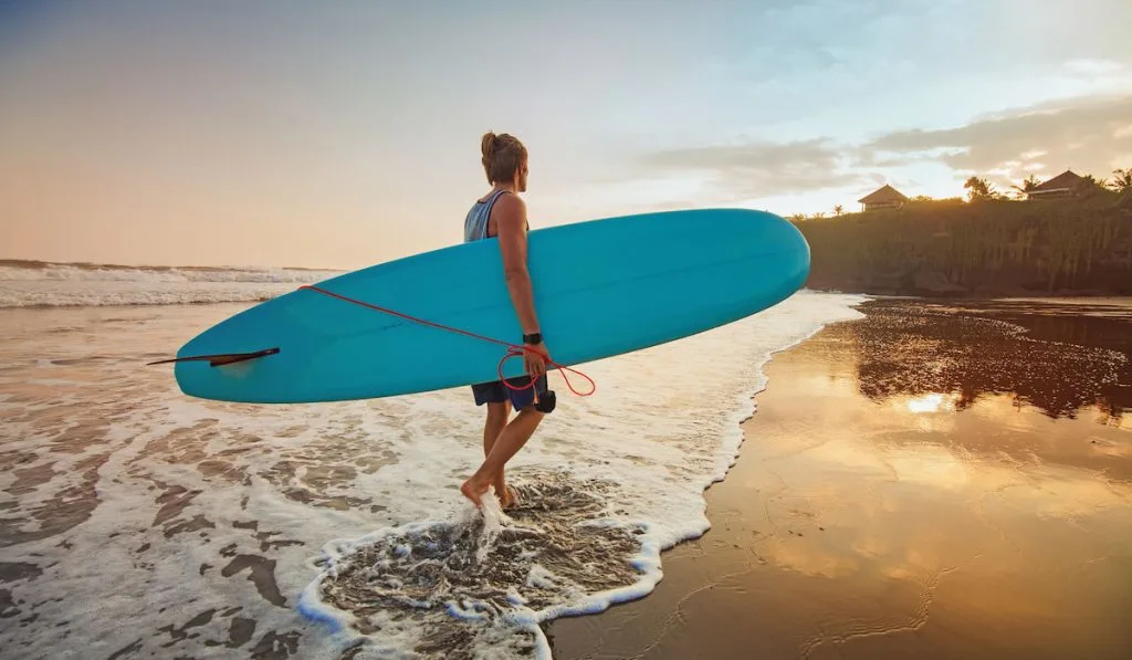 Guy walking on the beach holding his blue longboard for surfing