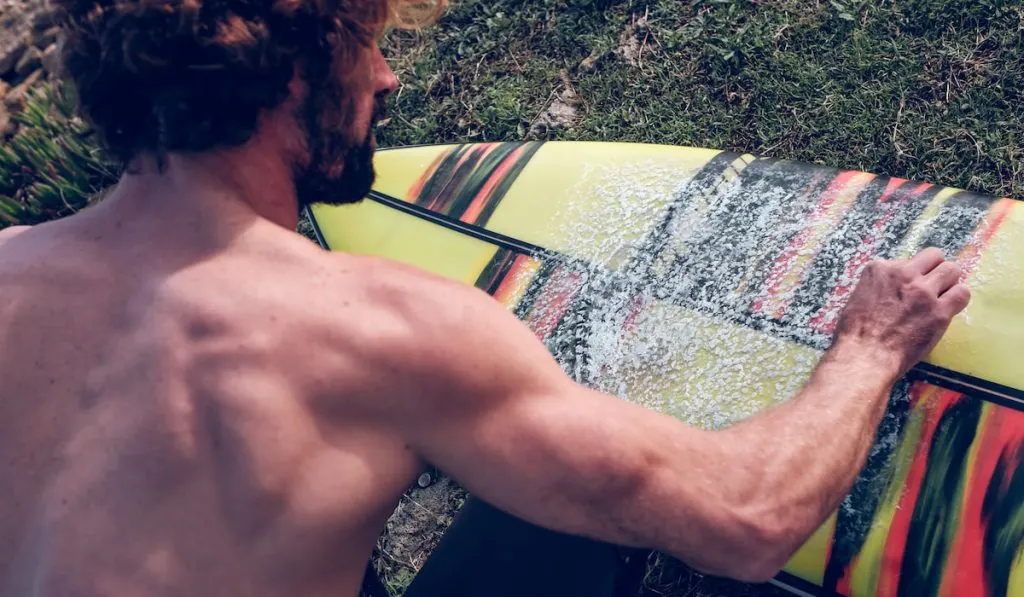 muscular male surfer covering surfboard with wax on grassy lawn